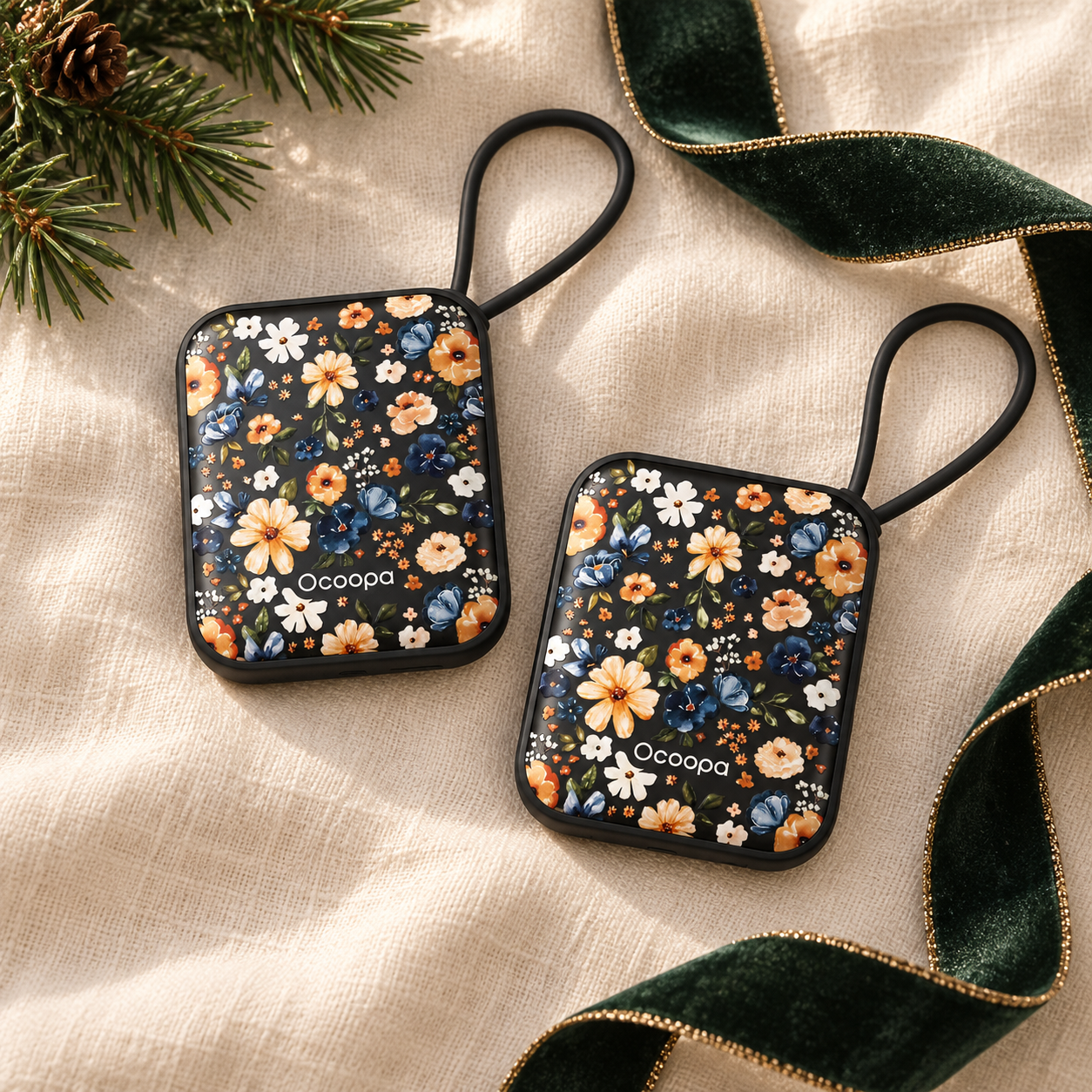 Two black floral Ocoopa hand warmers styled with an evergreen gift ribbon